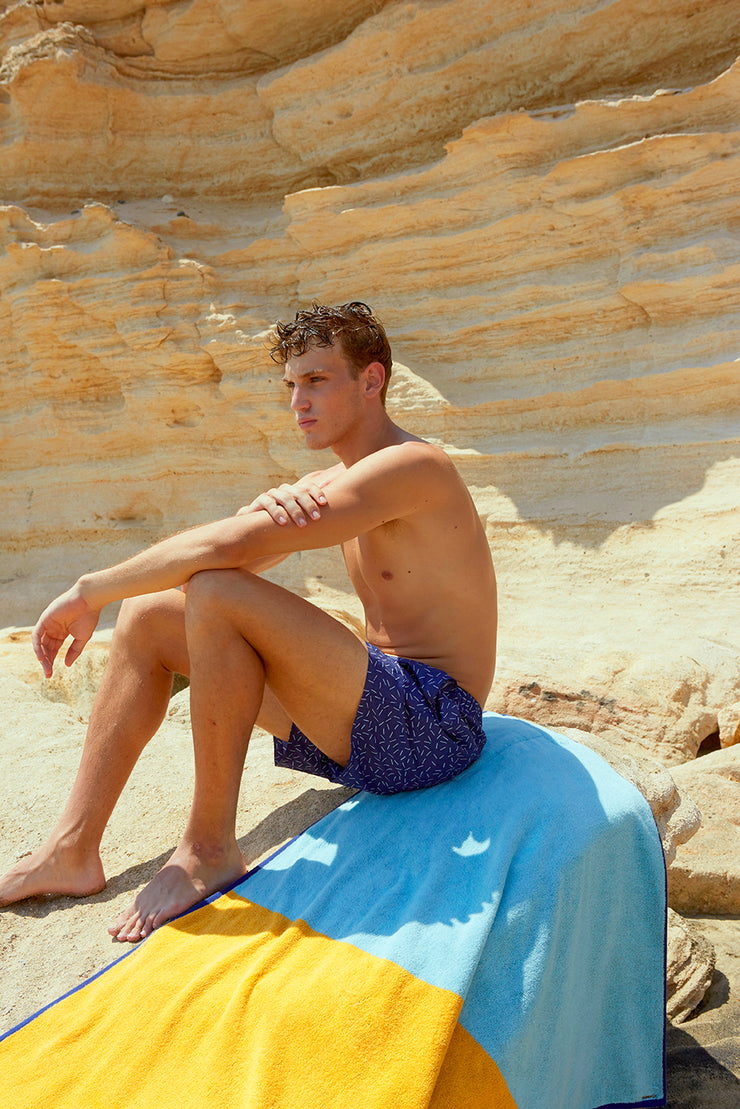 Hombre sentado en una roca sobre su toalla de playa Tucca "Dune". Azul claro, amarillo camello y azul oscuro en grandes bloques que componen un hermoso diseño. Una toalla de playa grande que no se vuela con el viento. Textura agradable y suave 100% de algodón.                                