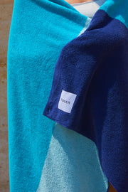 Chica arropándose con una toalla de playa Tucca "Swell". Colores azul claro, verde azulado y azul oscuro en en bloques de color dando lugar a un deseño bonito y elegante. Una toalla de playa grande que no se vuela con el viento. Textura suave y máxima absorción de agua. 100% algodón.