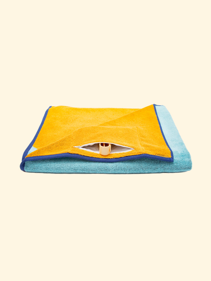 Modelo "Dune" de la toalla de playa de marca Tucca, doblada mostrando el bolsillo oculto impermeable con cierre de cremallera, que puede ser usado para guardar tu teléfono u otras pertenencias, así como para guardar tus pinzas Tucca después de usarlas para fijar tu toalla de playa a la arena.                                