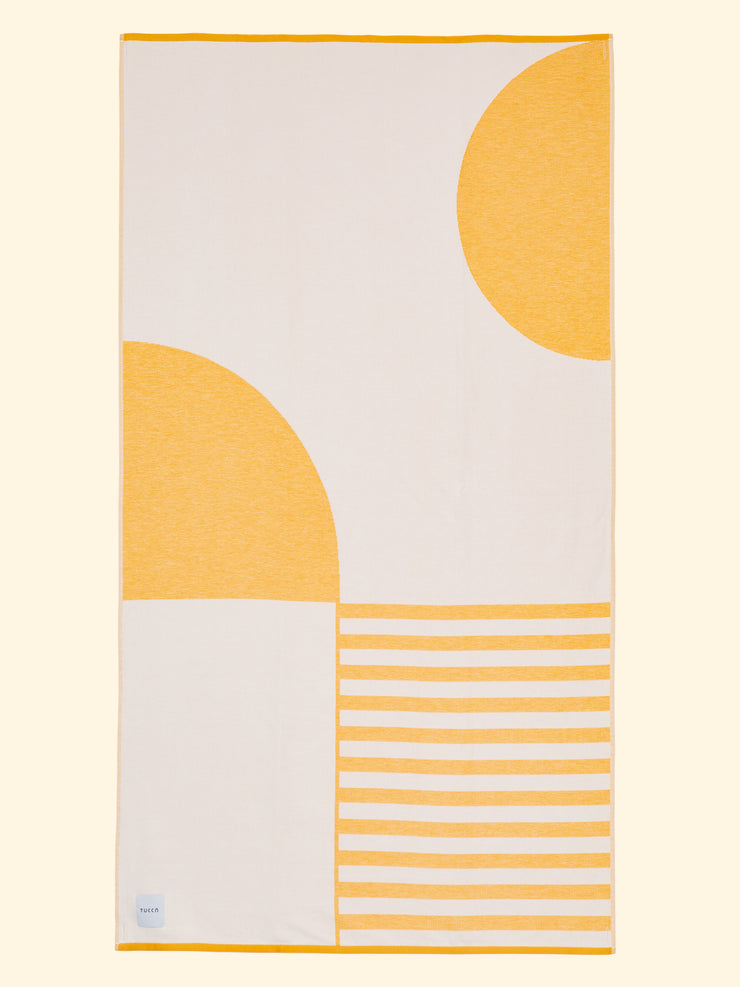 Tucca "Danai" toalla de playa extra ligera extendida. Colores amarillo mostaza claro y blanco en grandes bloques que componen un bonito diseño. Toalla de playa grande que no se la lleva el viento. Textura súper suave al estar fabricada con algodón 100% orgánico.