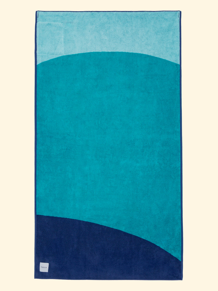 Toalla de playa Tucca "Swell" extendida. Colores azul claro, azul verde y azul oscuro en grandes bloques que componen un bonito diseño. Toalla de playa grande que no se la lleva el viento. Textura gruesa y suave al estar fabricada con algodón 100% orgánico.