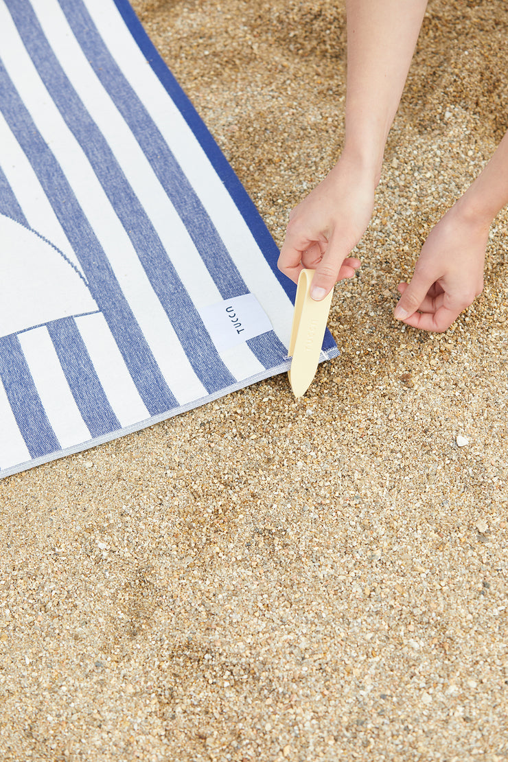 Imagen que muestra cómo fijar su toalla de playa en la arena para que no se la lleve el viento. Insertando el pasador Tucca a través de la esquina de la toalla de playa premium light estilo Florida. Fabricada en algodón 100% orgánico.
