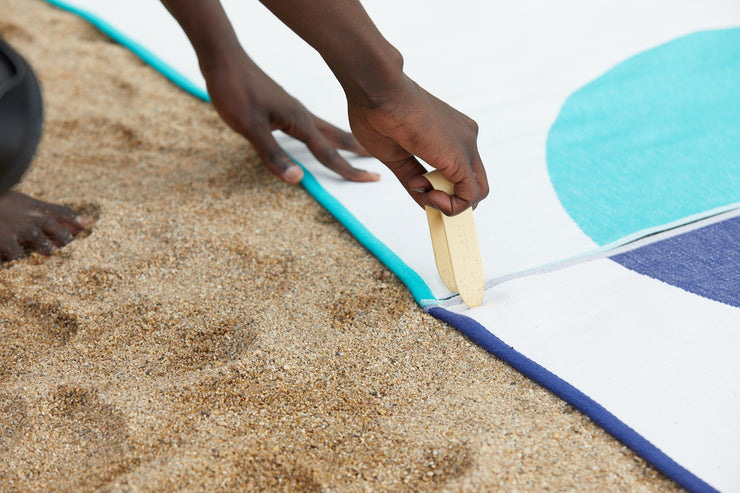 Picture showing how to fix your beach towels into the sand so they don't get blown away by the wind. Inserting the Tucca pin through the corners of the premium light beach towels Florida and Mayeri styles. This way both beach towels can be connected with each other creating a common design and a bigger space at the beach. Made of 100% organic cotton.