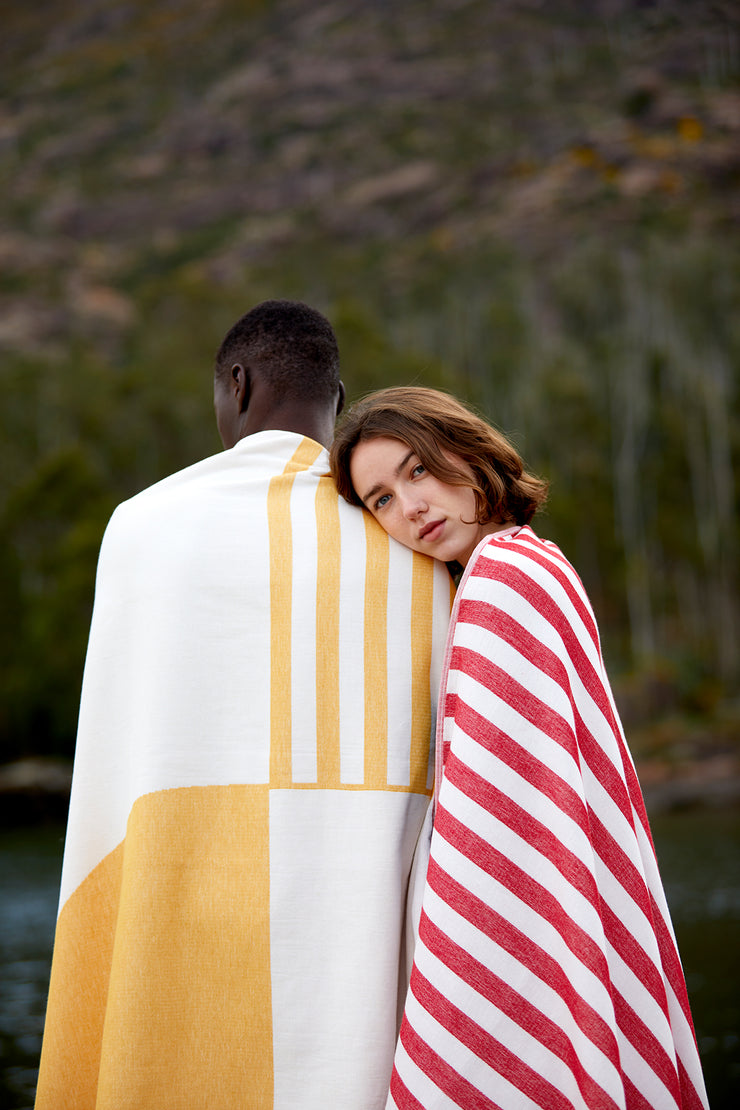 Una pareja se seca con las toallas de playa conectables Danai y Crassa. Ella se apoya en el hombro de él, ambos cubiertos con la toalla de playa premiums Tucca light de algodón orgánico.